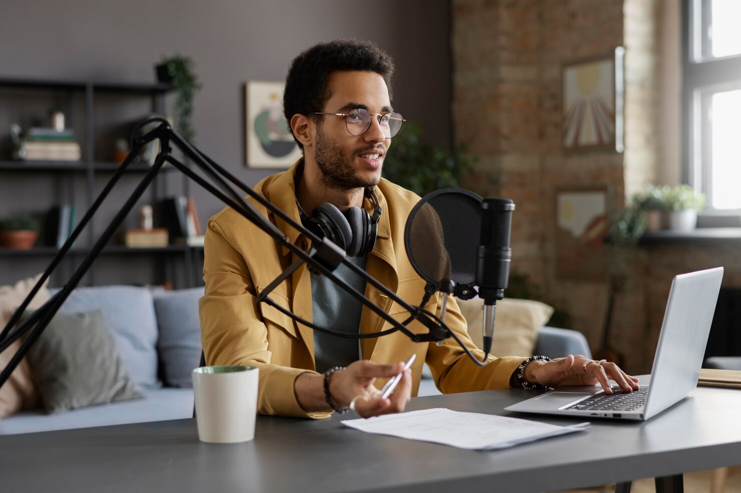 homem jovem e negro, com casaco amarelo, grava podcast em frente ao seu notebook com microfone profissinal