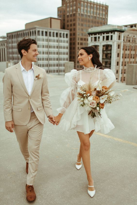 a groom standing with his wife wearing casual wedding dresses and holding a flower