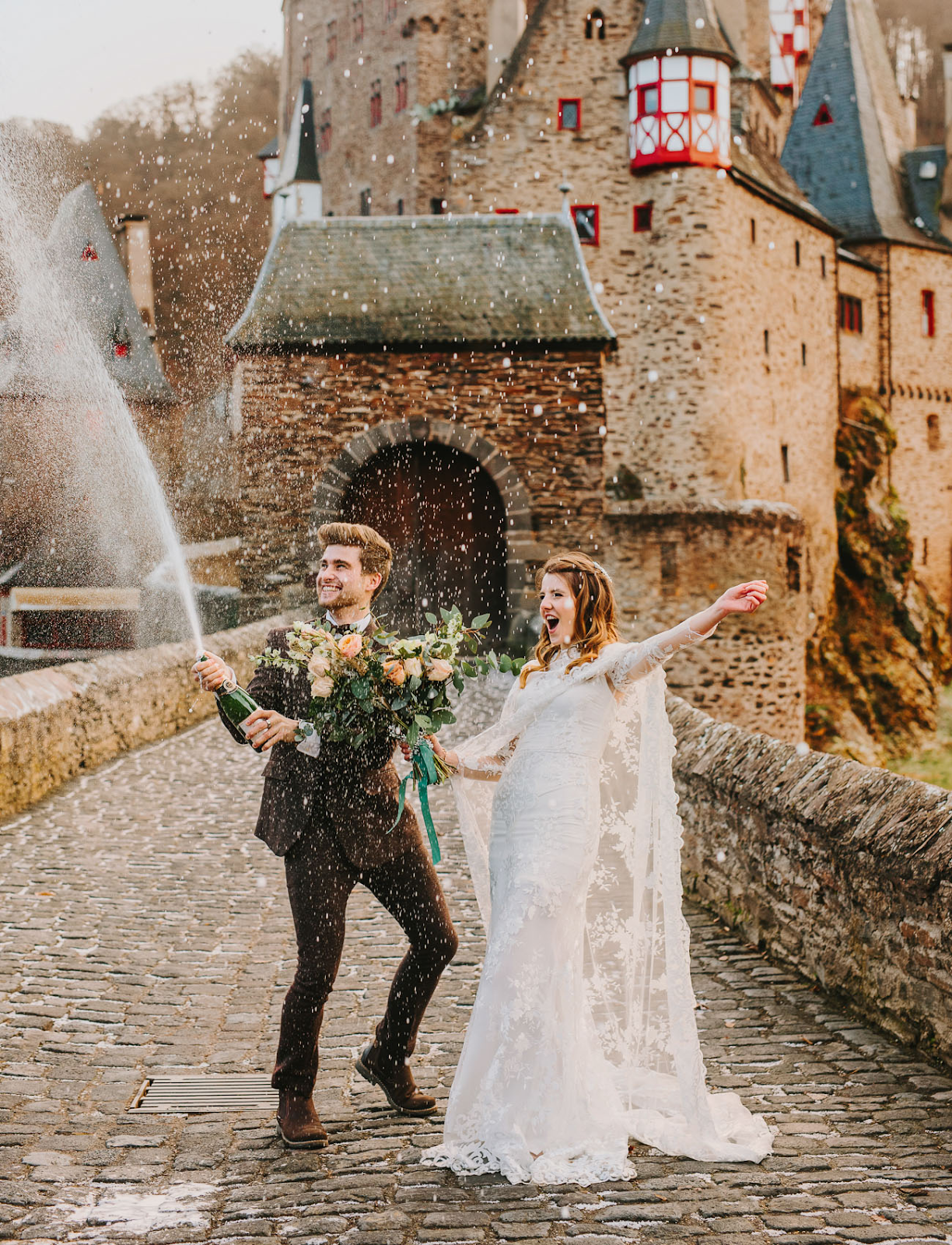 Casal de noivos em frente a um castelo antigo, abrindo uma garrafa de espumante em comemoração.