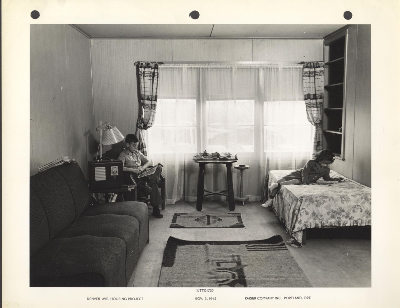 Black and white image depicting the interior living room. There is a couch, radio, lamp, a child reading in a chair, a table in front of a window, a child reading on a twin bed, and rugs on the floor. Typed on the image is: Interior, Denver Ave. Housing Project, Nov. 3, 1942, Kaiser Company Inc. Portland, Ore.