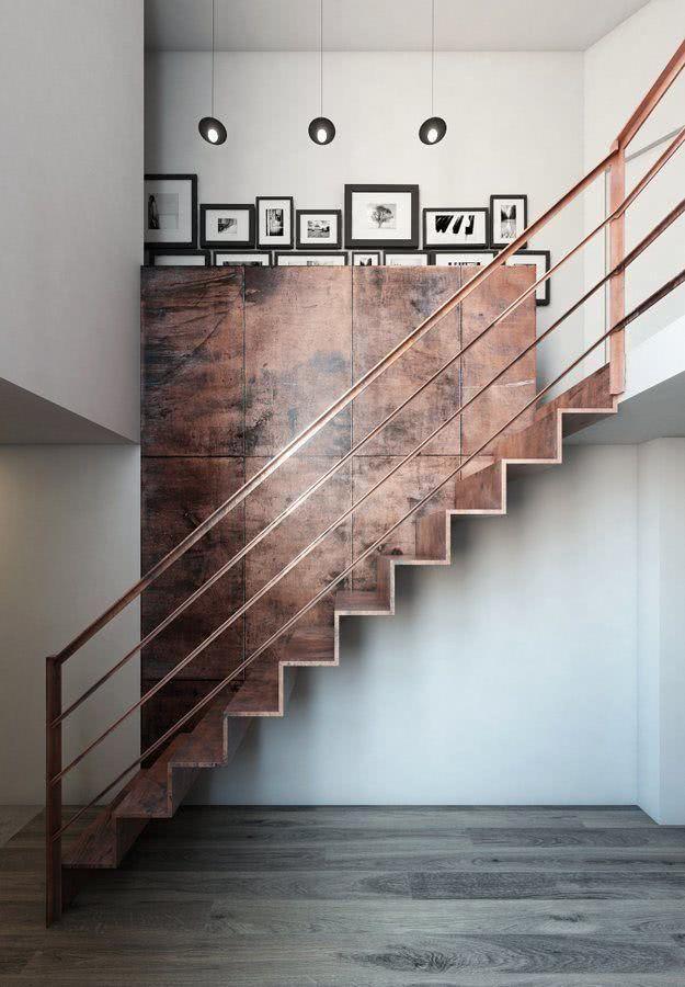 Ambiente com escada reta, pendente na parede, na cor cobre com corrimão na cor cobre e revestimento na parede ao lado amadeirado.