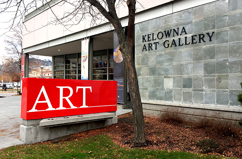 An outside view of the Kelowna Art Gallery with a brick wall and red sign that reads ART