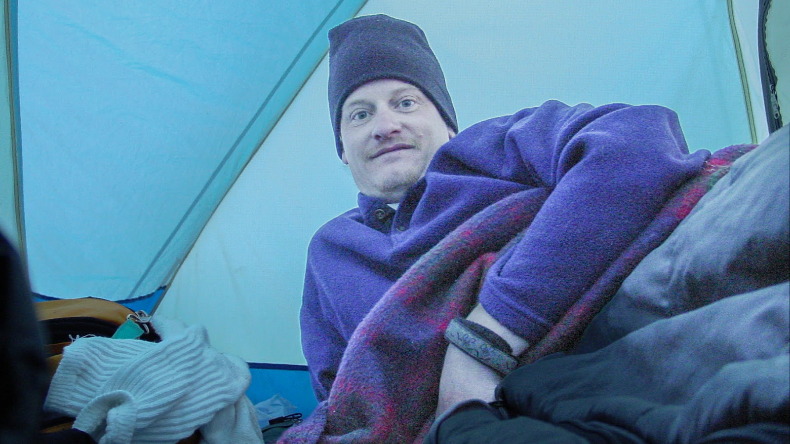 A man looks up from a sleeping bag in a crowded tent.
