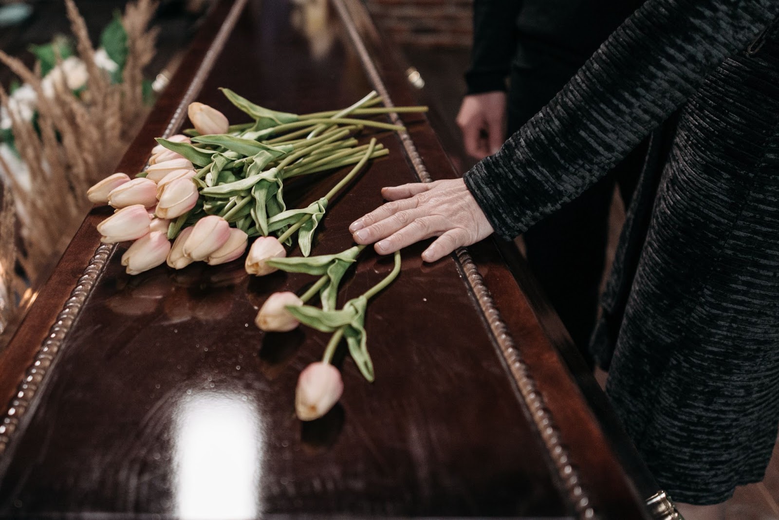 na foto vemos um caixão com algumas flores em cima, uma mulher está encostando sua mão em cima da urna funerária