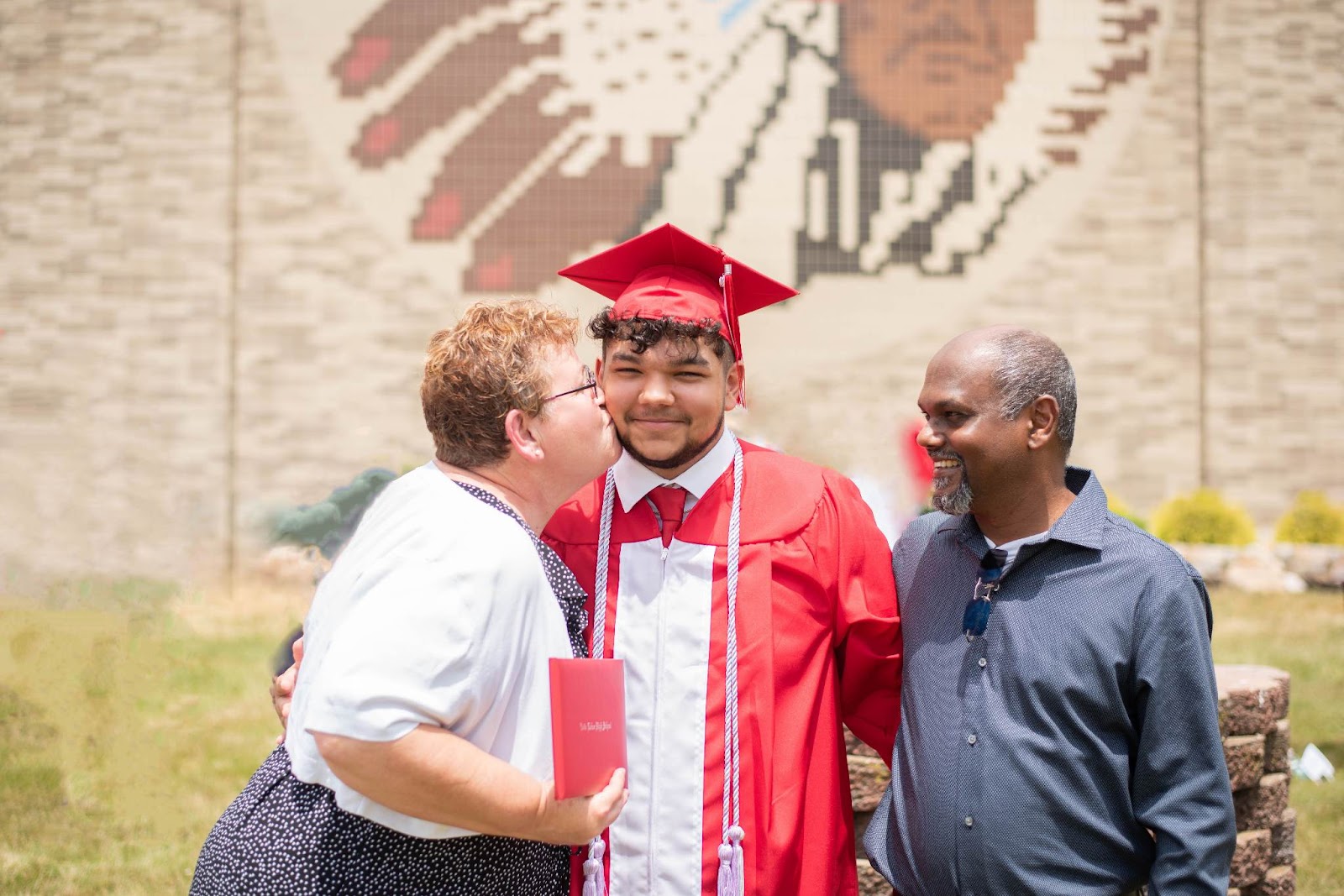 Bryce Beharry standing with his mother and father outside of his high school wearing his cap and gown, all smiling with his mother kissing his cheek