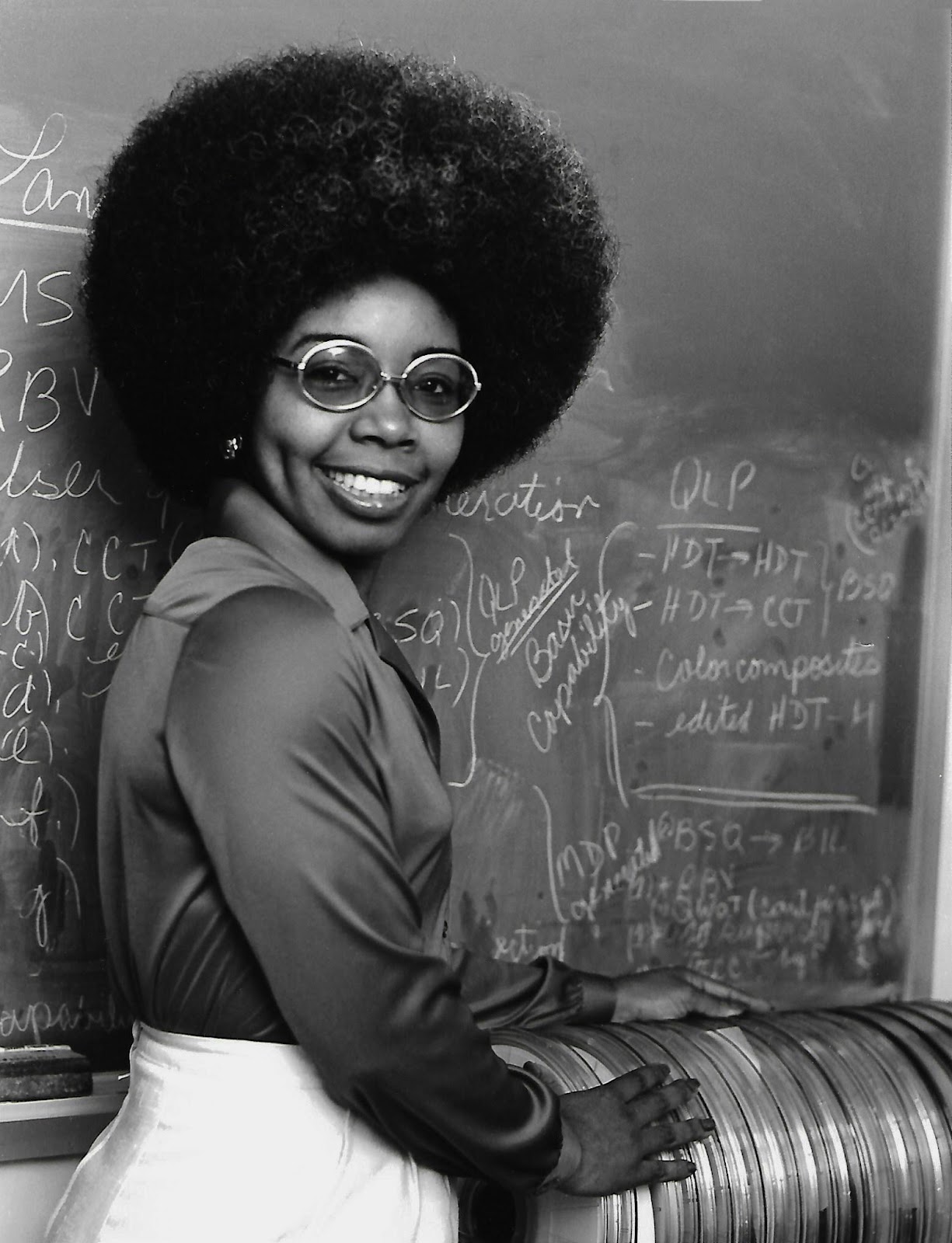 Black and white photo of Valerie Thomas posing in front of a chalkboard covered in notes and calculations. Thomas is styling an afro with round tinted glasses, a dark-colored blouse and white bottoms.
