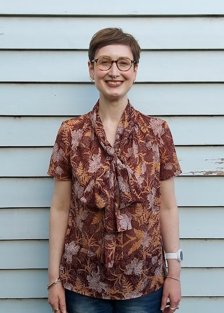 Siobhan stands in front of a blue weatherboard wall. She wears an earthy brown, fern-print short sleeve blouse with comically large bow at neck. She is smiling.