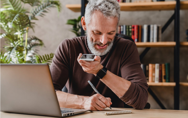 An older man with silver hair and a beard, sitting at his laptop while speaking on his mobile phone and taking notes on a notepad