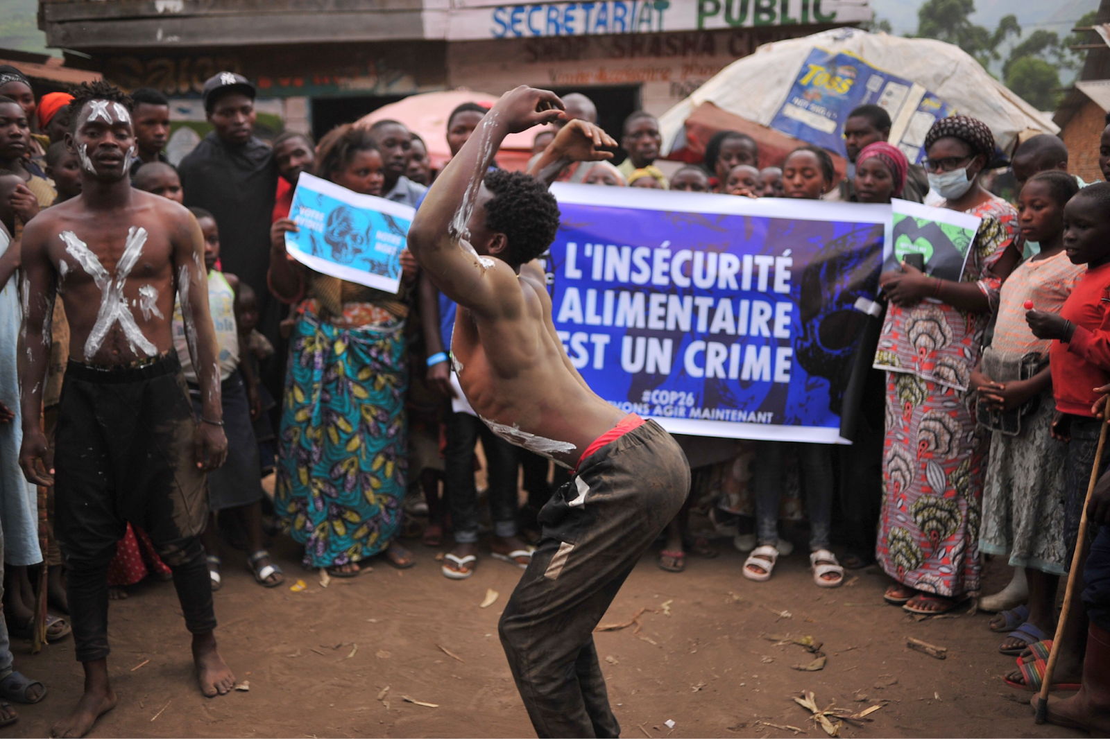 Two Congolese rebels dance with white paint daubed over their faces and chests. Villagers surround them.