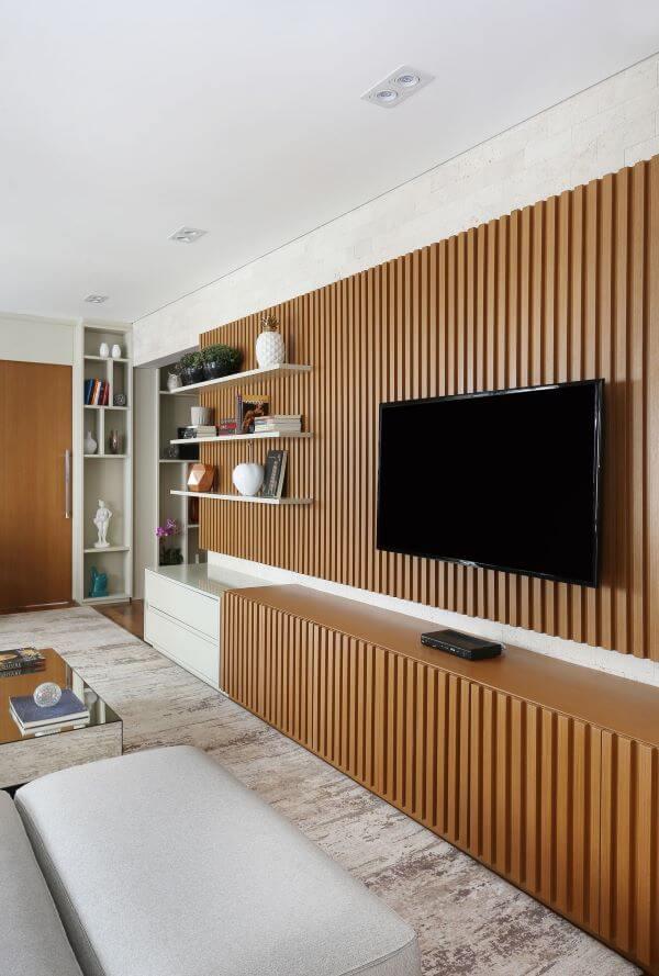 Ambiente com painel de TV e rack de madeira ripada, móvel branco com prateleiras brancas, tapete e poltronas claras.