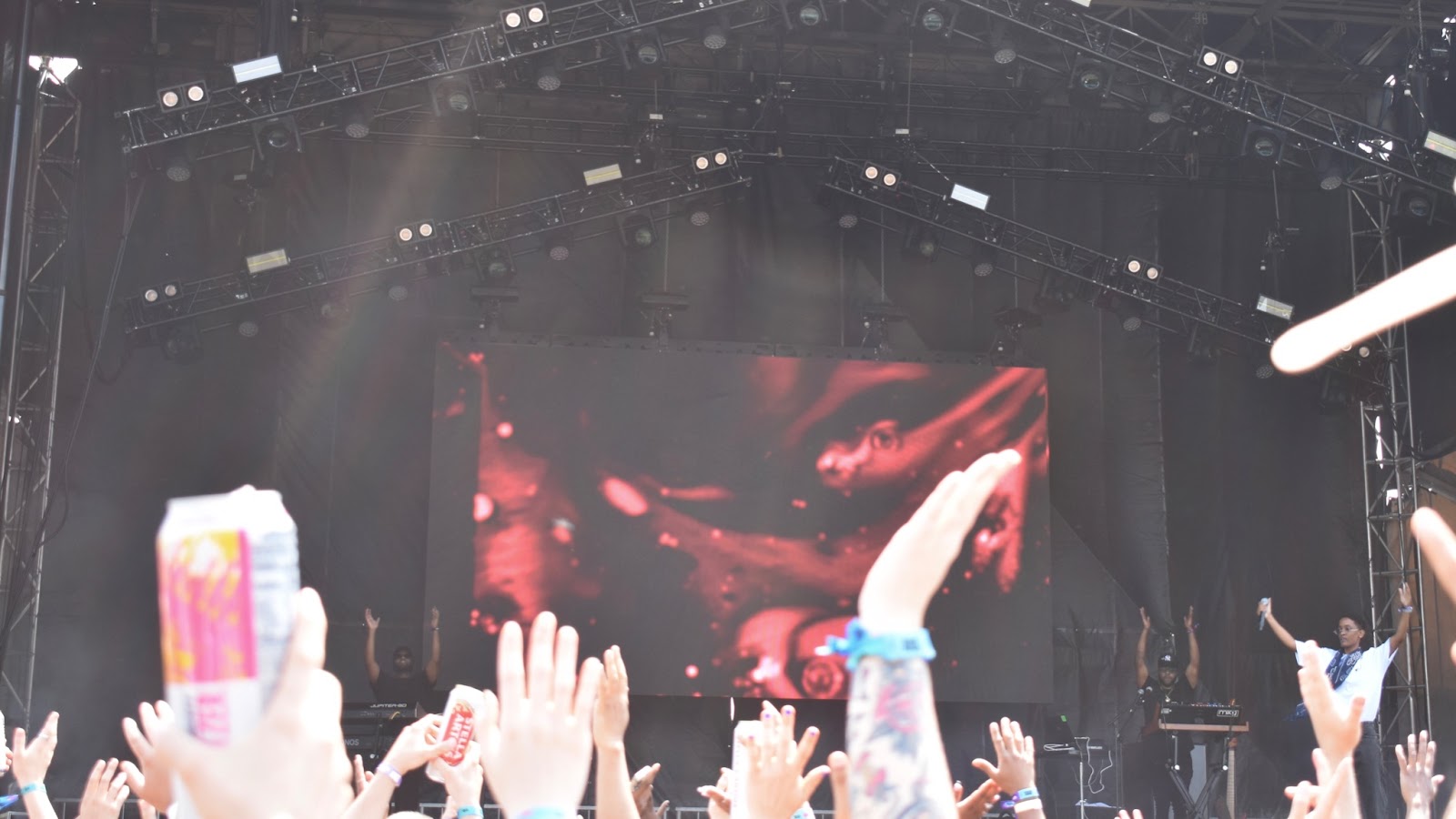 Hands waving in the air in front of a red backdrop at a daytime concert.