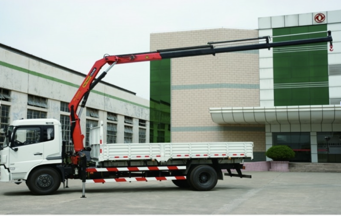 A truck mounted crane with white car paint, and the truck's crane is mounted.  The truck mounted crane parks in front of two buildings. Those buildings have white bricks and green glasses.