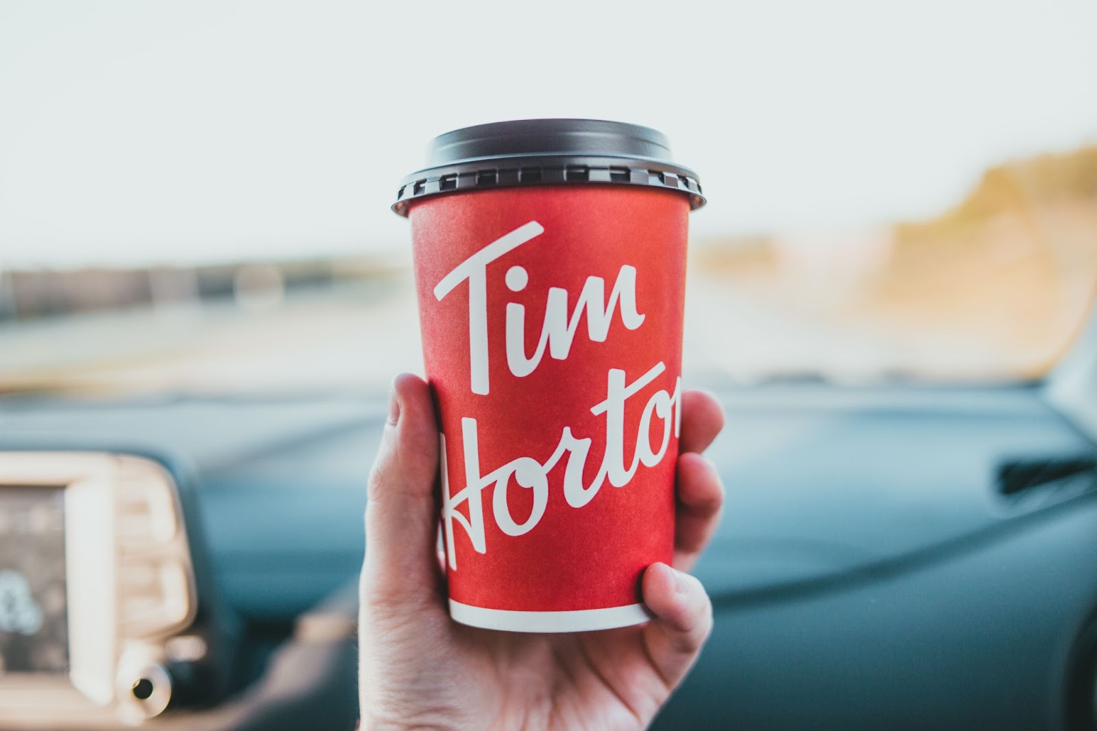 a hand holding a Tim Hortons red cup, with a car dashboard in the background.