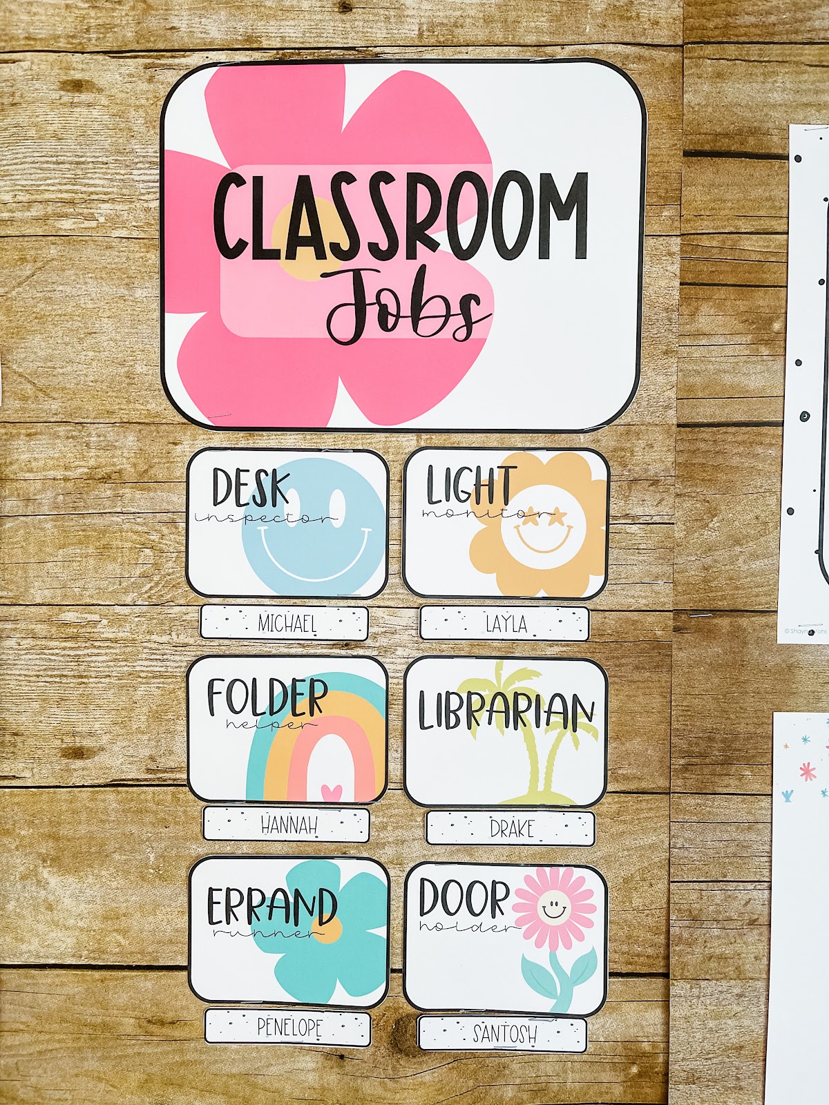 This image shows a classroom jobs display. There is a large card labeled "classroom jobs" with a flower in the background. Underneath that larger card are smaller cards with the names of different classroom jobs on them. For example, "Desk inspector" or "Door holder". Under each job card is a small rectangle with a student's name. The cards all have large images in their background to create a tropical, beach theme. 