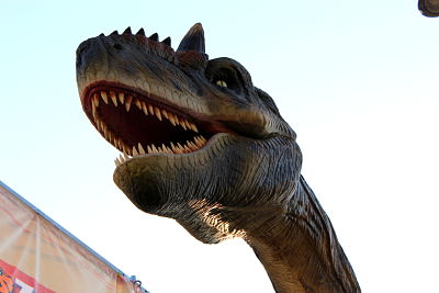 Cabeza y cuello alargado de una réplica de tiranosaurio rex con colmillos afilados, dispares y ensangrentados, arrugas en la piel verdosa con matices cafés y blancos, un cuerno en medio de los ojos amarillos y seis puntas sobre su hocico, con el cielo de fondo.
