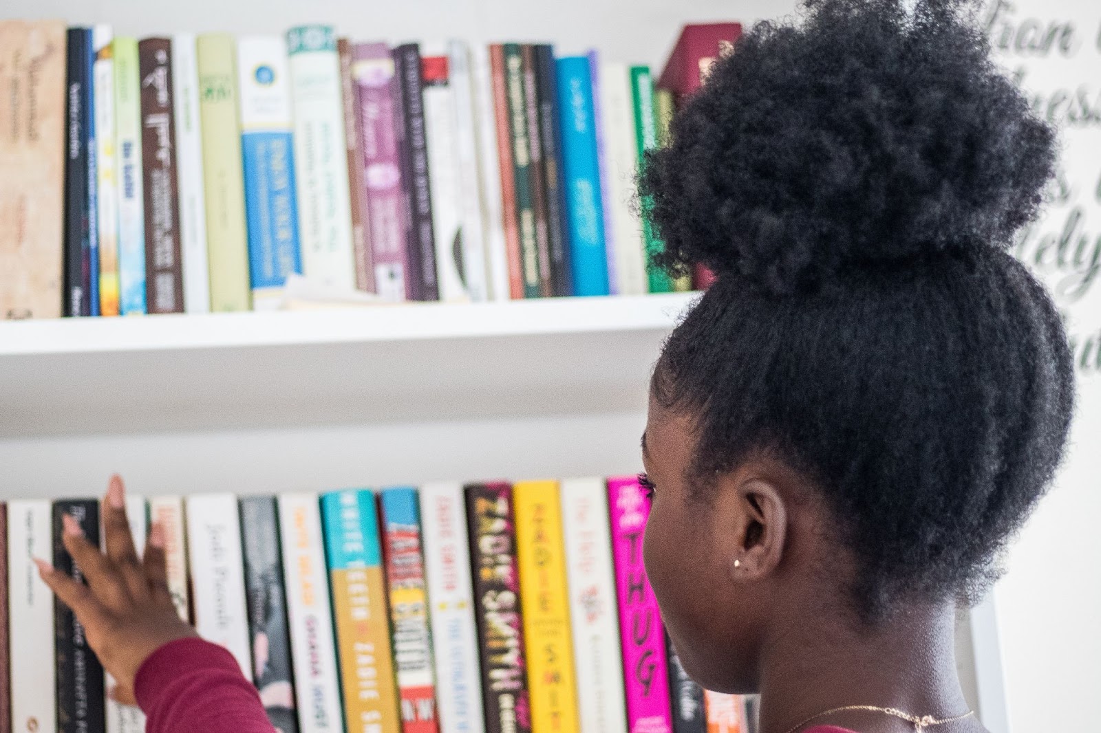 Young Black woman with high ponytail touches books on a shelf.