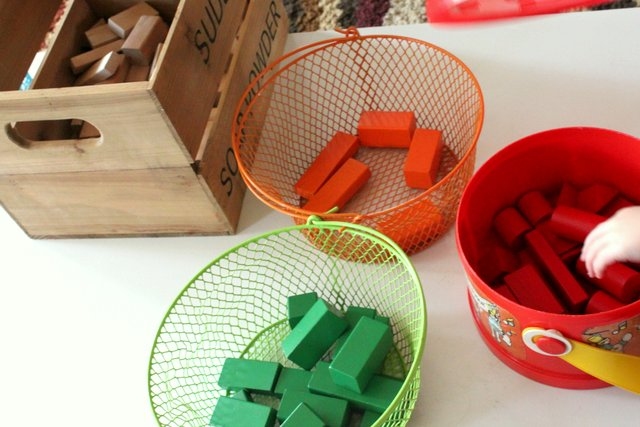 A wooden crate with rectangular wooden blocks sits next to an orange wire basket with several orange wooden blocks. There is also a green wire basket full of green wooden blocks in the foreground of the photo. To the right is a round red plastic container with red wooden blocks. A child's hand reaches into the red container to grab a block.
