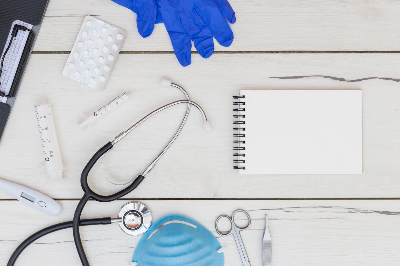 Medical equipment, gloves, pills, and spiral notepad on a white wooden desk, representing the essential tools for studying medicine in the UK.