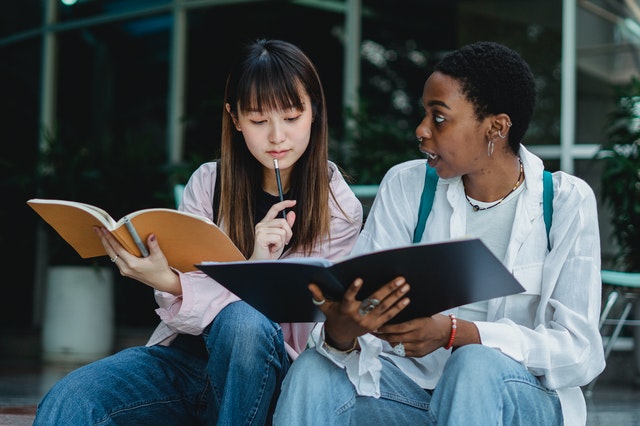 Two young women study side by side, one consulting a book while the other tells her something.