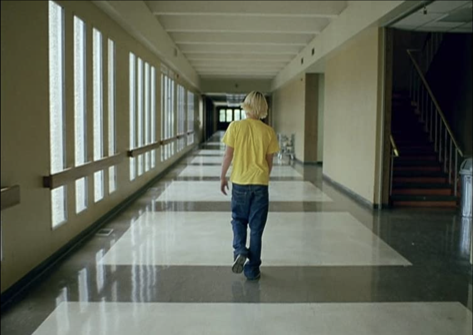 A blonde-haired person wearing a yellow t-shirt, baggy jeans, and sneakers. They walk down a school hallway, seen from behind at a distance.
