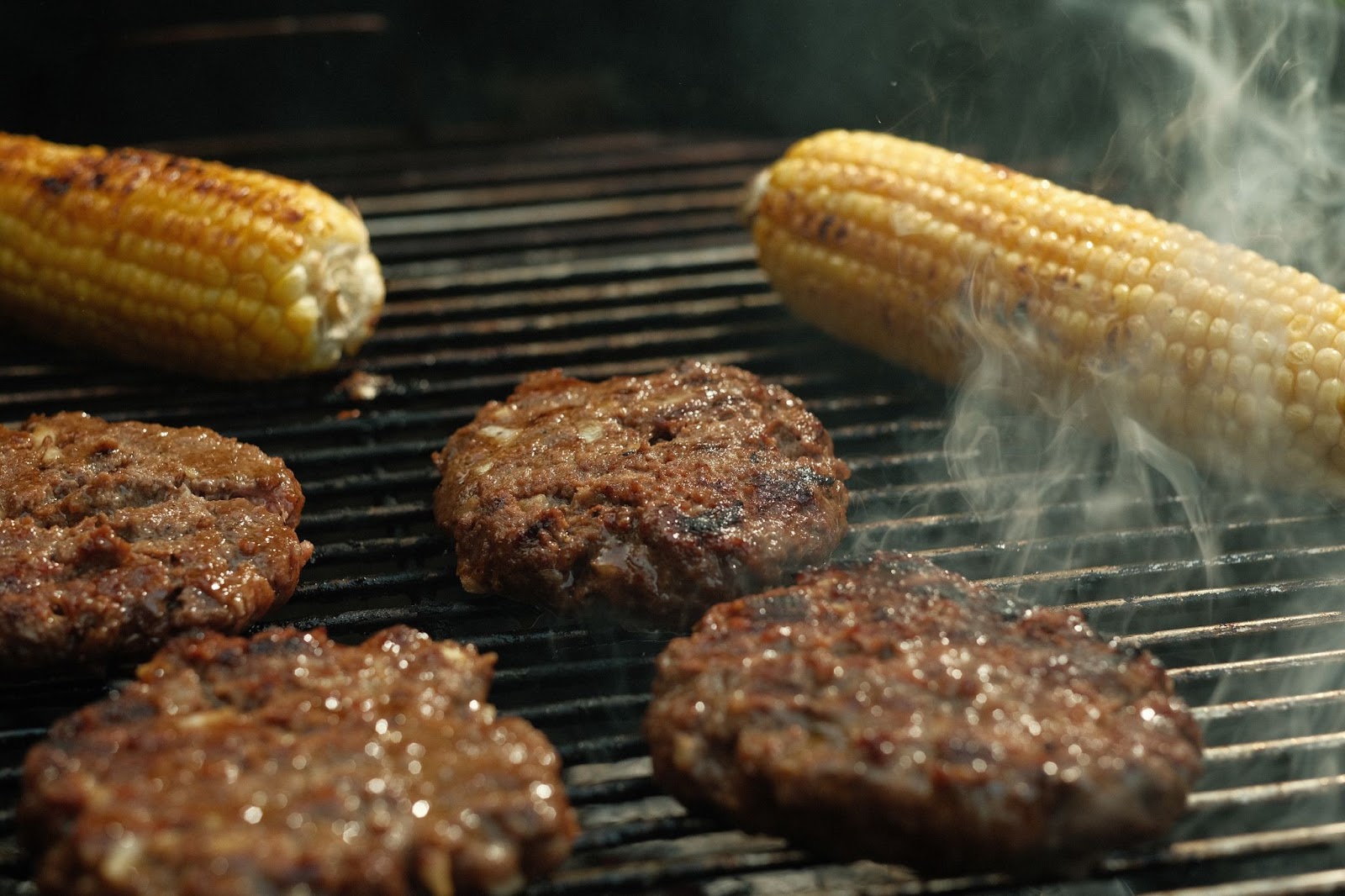 four burger patties from T Bones in Kelowna, on the grill smoking with lightly charred corn on the grill in the background
