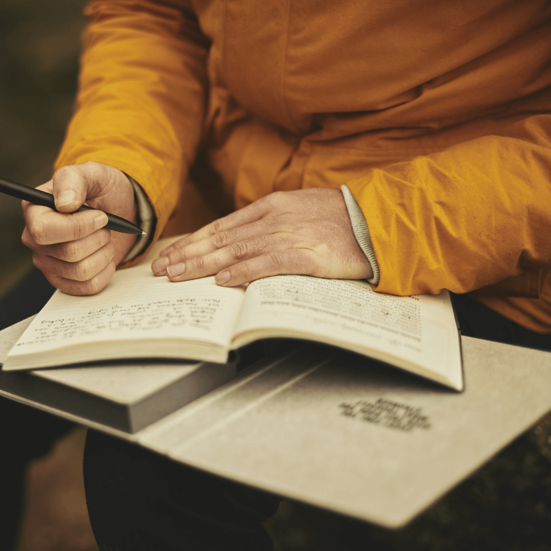 The image shows a person sitting down and writing in a journal. The image is zoomed in where you can only see the hands, journal and a portion of the person's yellow jacket. The image is focused on the individual's hands where they are pausing while writing, the rest of the image is out of focus. 