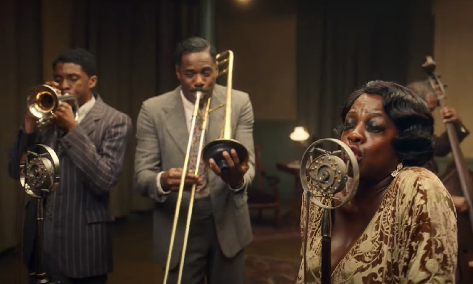 This is a screen still from Ma Raineys Black Bottom. Viola Davis is singing into a microphone while in the background, two men play brass instruments.