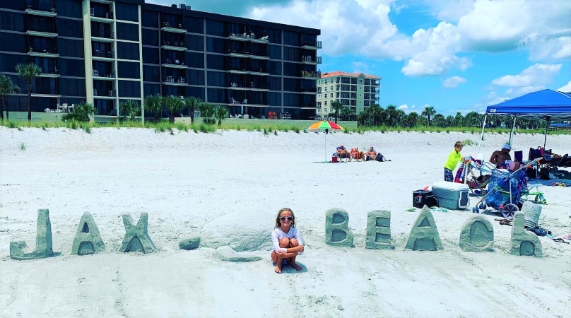 A child crouches down and smiles on the beach next to her sand creations that spell “JAX BEACH”