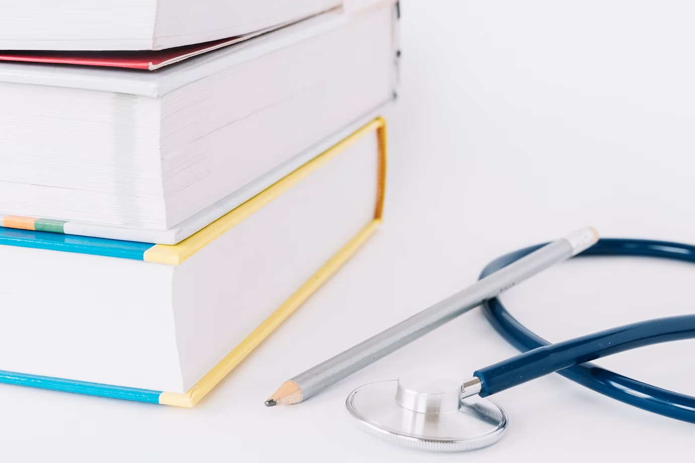 Stacked books, pencil, and stethoscope on a white surface – Top pre-med subjects for a medicine career.