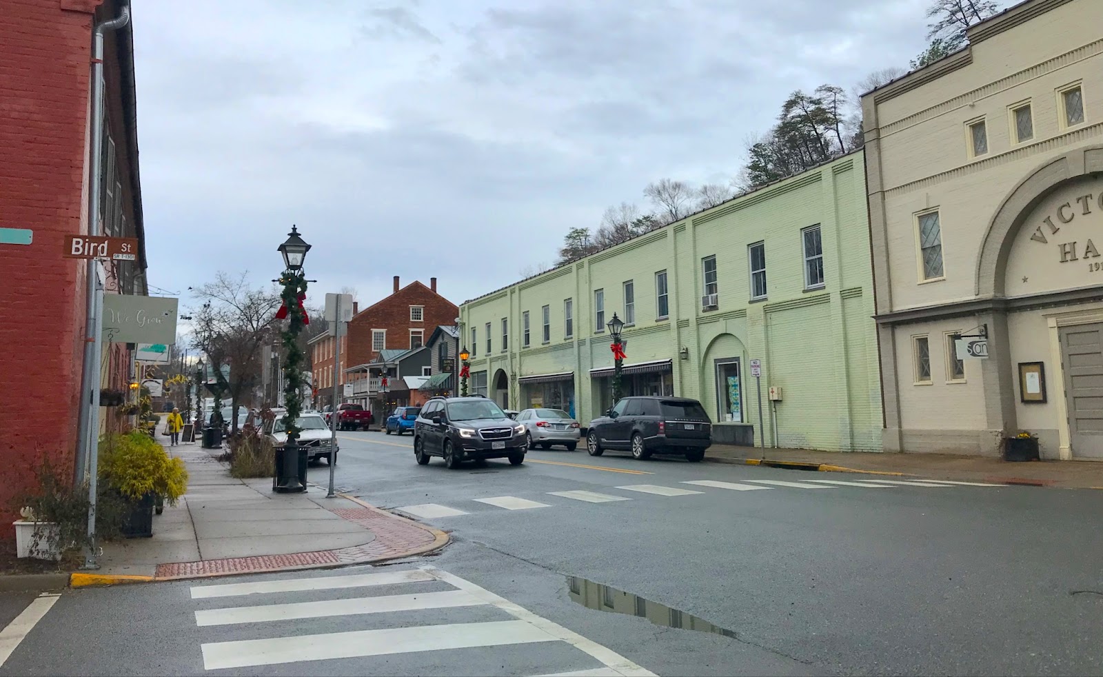 A two-lane street with parking on both sides runs through the middle of a small town, with older brick buildings painted different colors. The lampposts and bare trees are decorated for the winter holidays.