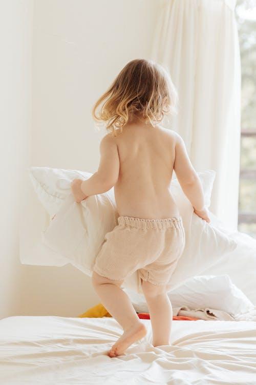 Free Back view of anonymous little girl in casual clothes playing with pillows on comfortable bed in light room at home Stock Photo