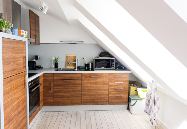 A small attic that’s been renovated into an extra kitchen space with a stove top, oven, sink and wooden cabinets. The white walls contrast beautifully with the natural wood cabinetry.