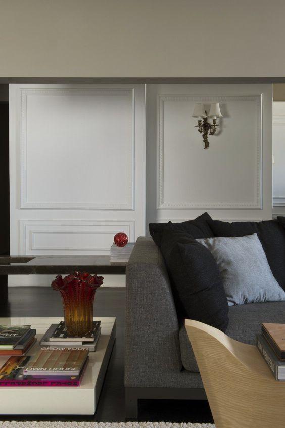 Sala com sofá cinza e almofadas pretas, parede branca com boiserie e mesinha de canto branca com livros e vasos.