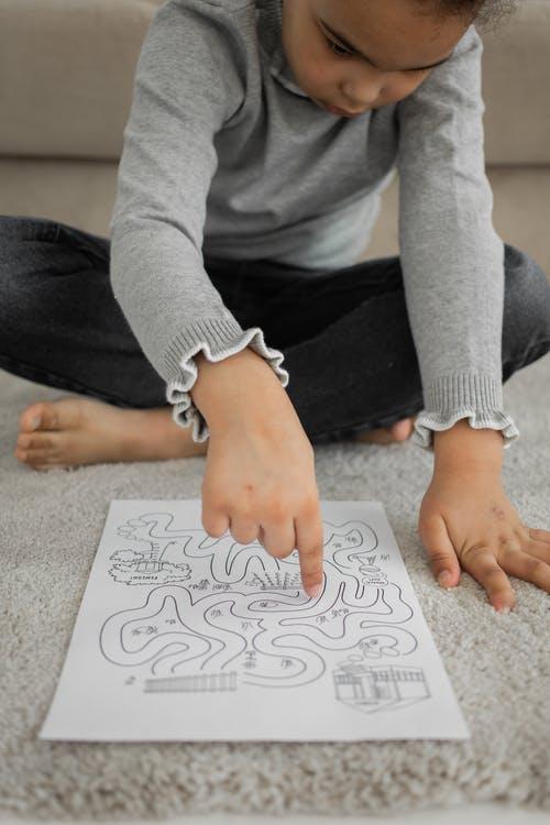 From above of crop attentive little black girl in casual clothes passing path of logic labyrinth game with finger sitting on floor at home