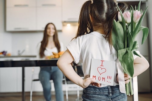 Dziewczynka chowa za plecami kwiaty i laurkę z napisem "I love mam". W tle rozmazana, uśmiechnięta mama.