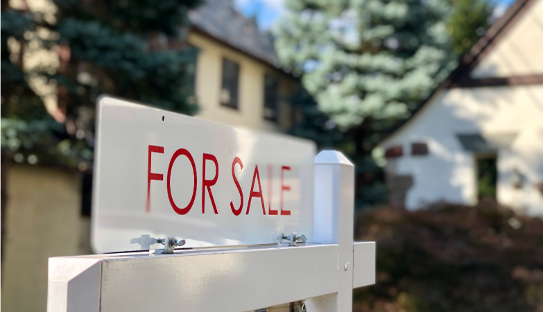 A “FOR SALE” sign in front of a residential home among pine trees.