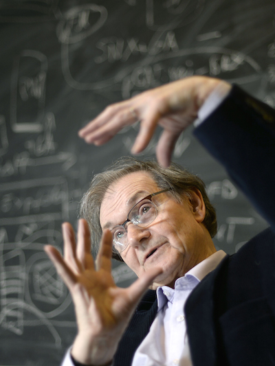 Sir Roger Penrose from the mid-torso & up gesticulating in front of a filled chalkboard.