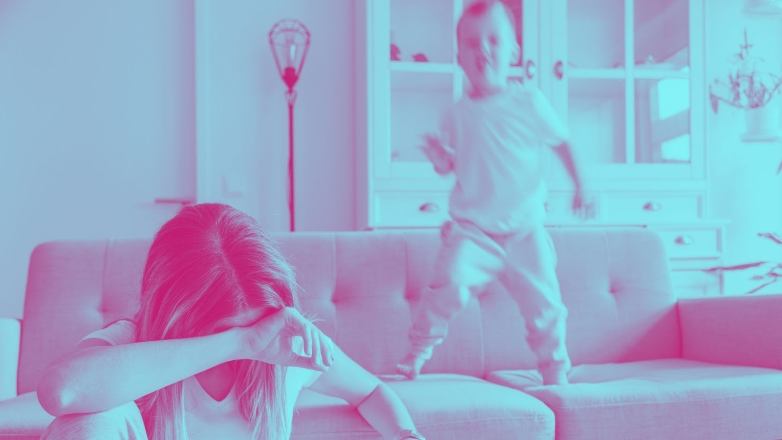 Fotografia colorida com filtro bicolor rosa e azul, mostrando um menino de 5 anos pulando ativamente no sofá, com sua imagem borrada pelo movimento, e uma mulher branca, de cabelos loiros, cobrindo o rosto em posição de desânimo, sentada na frente do sofá. Ao fundo, está uma estante de madeira branca e vidro
