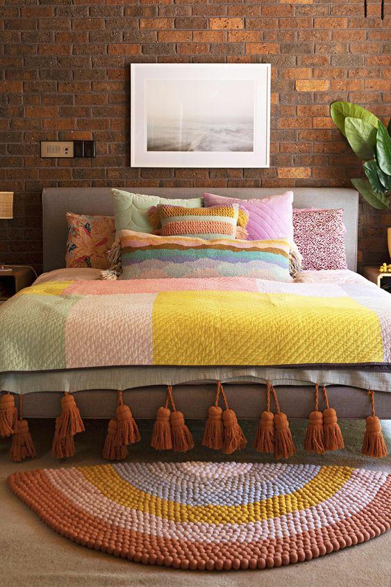 Quarto de casal rústico com tijolinhos na parede, as roupas de cama coloridas e o tapete colorido, cabeceira estofada cinza e vaso de planta no criado mudo.