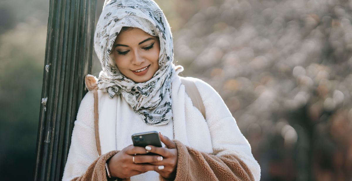 زبونة مسلمة محجبة مبتسمة تقرأ رسالة مبهجة في رمضان على هاتفها الذكي