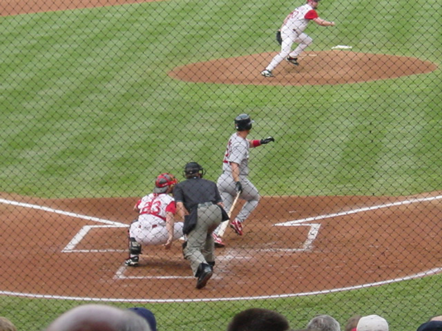 The batter twists at home plate watching the ball he hit as the pitcher reacts moving to the right side of the frame.