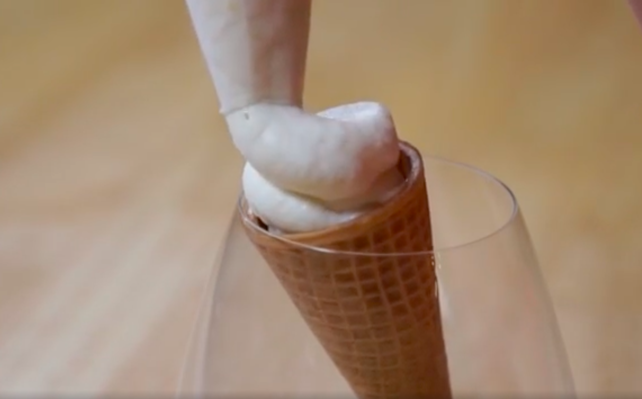 White soft serve ice cream piped from a piping bag into a sugar cone stood up in a wine glass. 