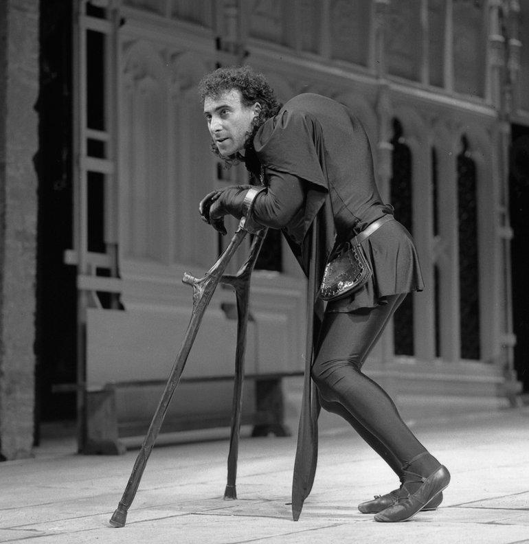 Black and white photograph of an actor, dressed as a medieval prince, leaning on crutches and with an exaggerated hump on his back.