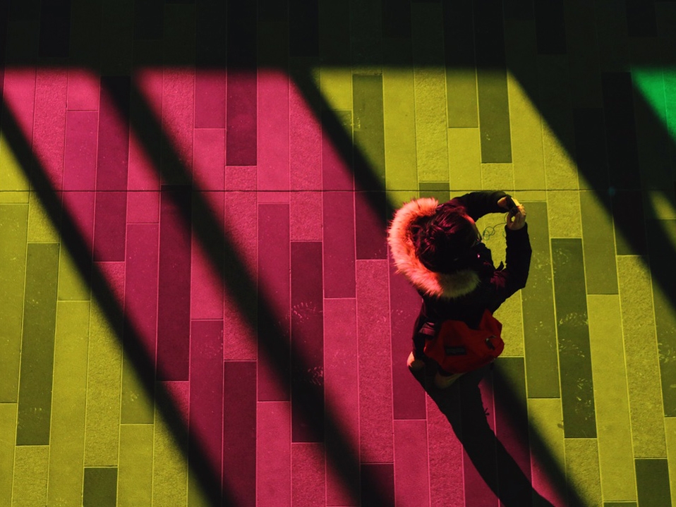 An aerial shot of a girl in a coat holding a cell phone up to her face. She&#039;s standing on a floor that&#039;s pink, yellow, and green from the lights shining down on it.