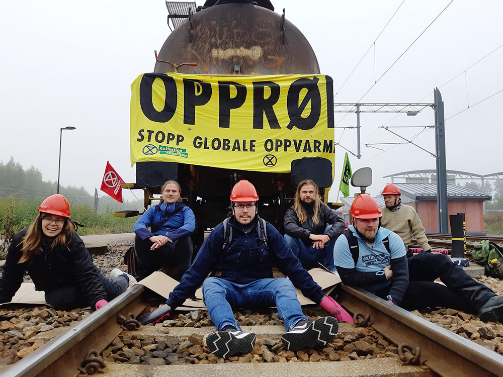 Rebels, some in red hard hats, sit locked to rail tracks in front of a rail tanker covered with a banner saying Stop Global Heating in Norwegian