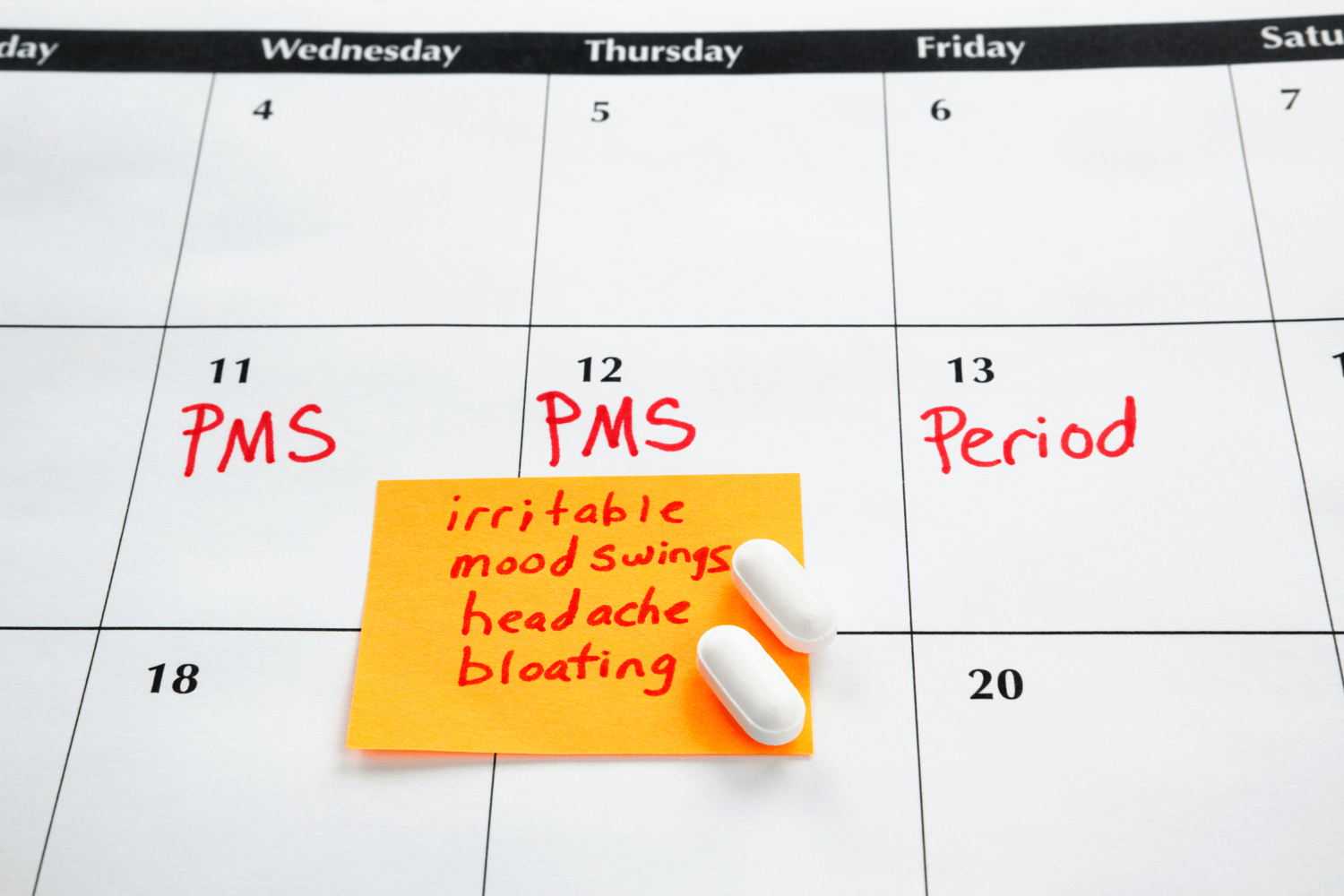 Calendar with PMS dates notated and PMS symptoms on a post-it note with 2 white pills sitting on top