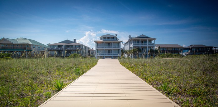 A long, wooden boardwalk leads to a vacation house rental at a beach.