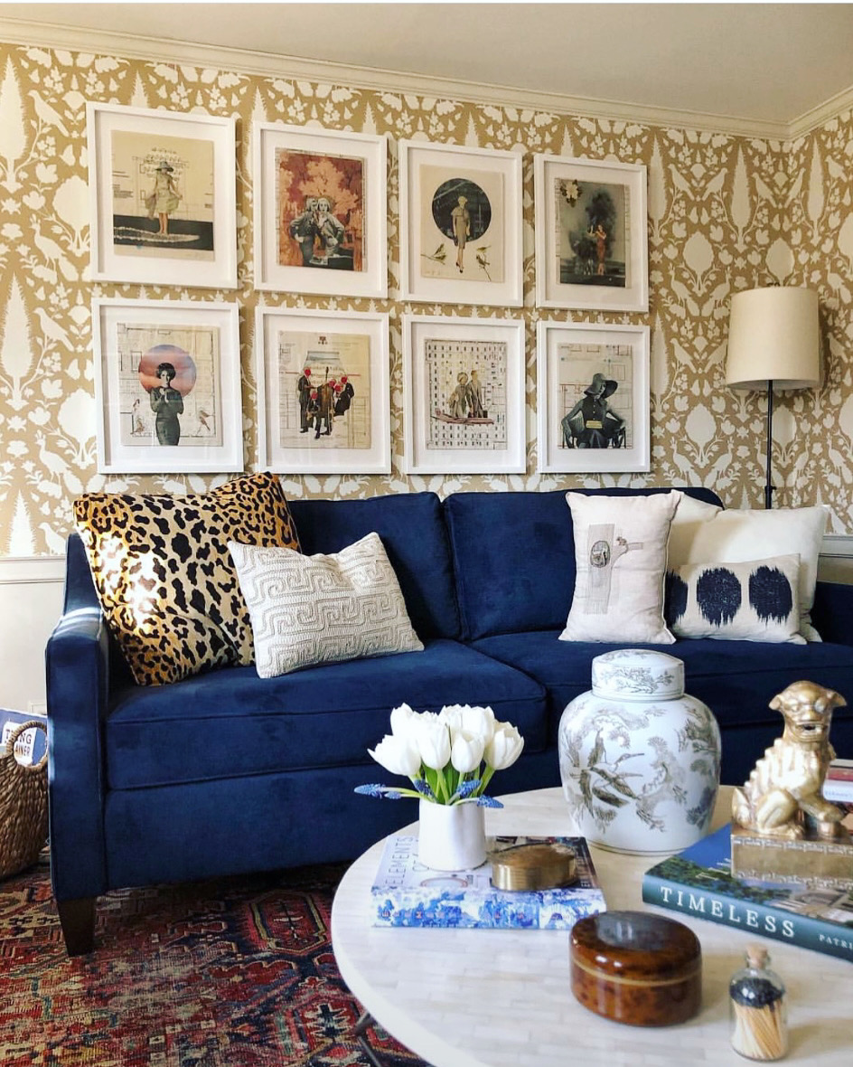 A bold living room features a cobalt blue velvet sofa, traditional round marble-top coffee table, large-print wallpaper and a variety of decorative accessories that pull the look together.