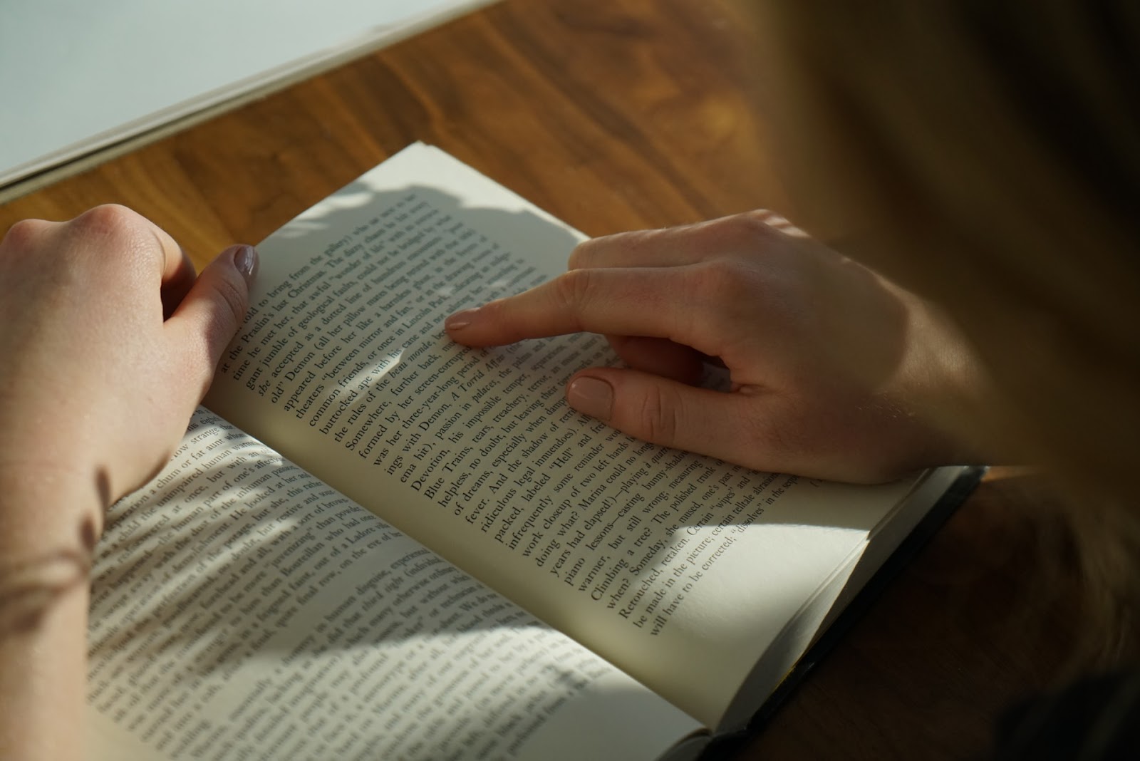 A imagem mostra as mãos de uma mulher sobre um livro. Com o dedo indicador da mão direita ela acompanha a leitura do livro. Também é possível ver os cabelos loiros da mulher desfocados no canto direito da imagem. 