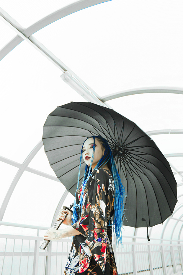 Well-lit image of a woman with blue dreadlocks and bold makeup holding a black parasol. She is wearing a serious expression and turned slightly toward the camera.